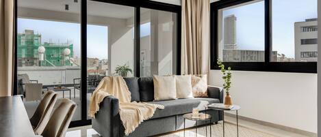 Un salon moderne avec un canapé gris, une table basse et une vue sur le paysage urbain grâce à de grandes fenêtres.