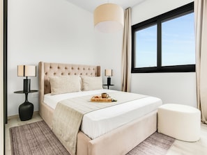 A modern bedroom with a tufted headboard, two bedside lamps, a bedside table, a window with curtains, and a small ottoman. - Sanders Premier Downtown - Full Building (Limassol)