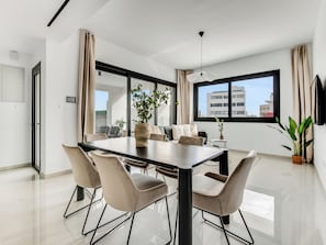 A modern living room with a dining table, beige chairs, a potted plant, and a large window with a city view. - Sanders Premier Downtown - Full Building (Limassol)