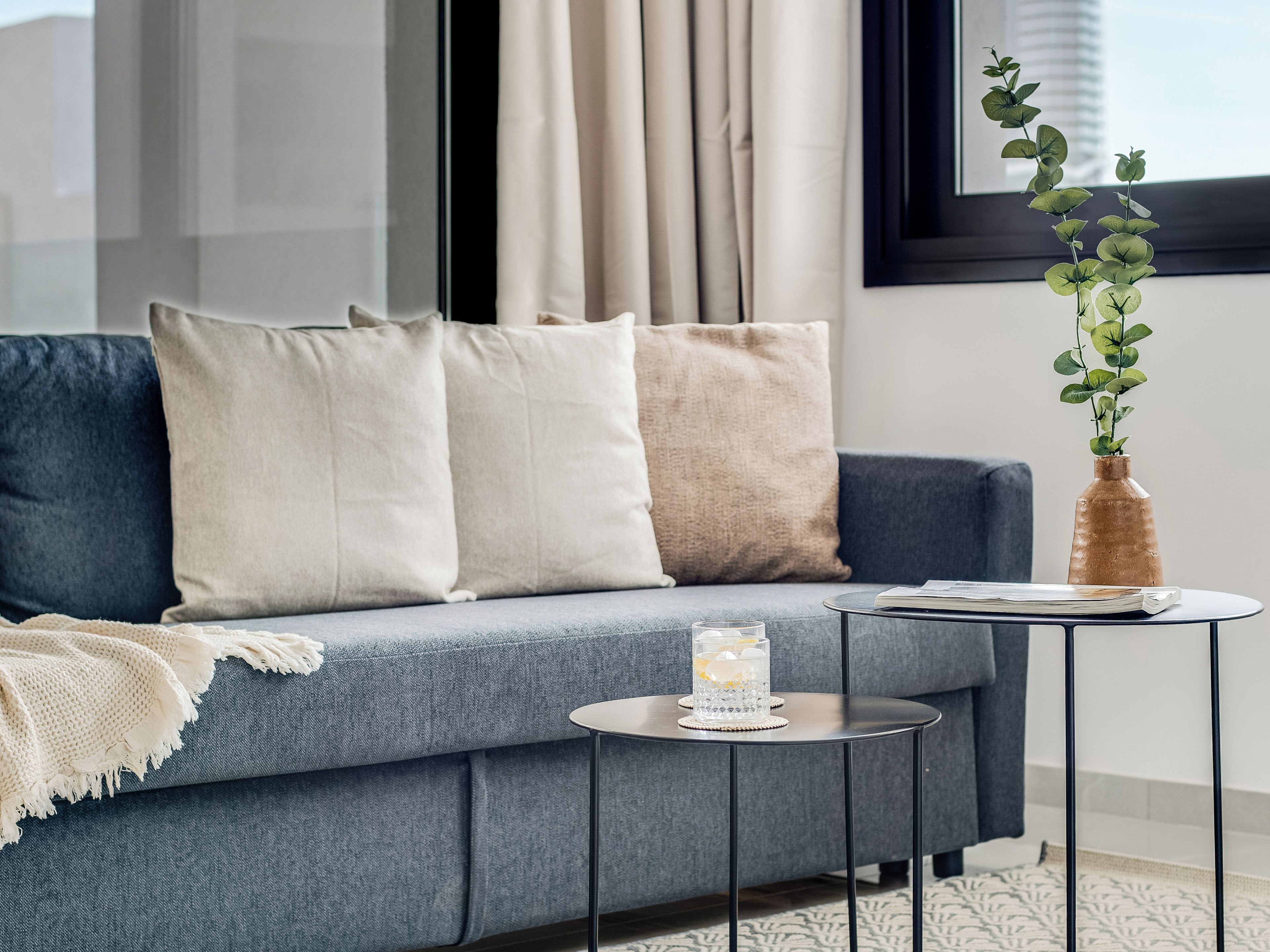 Un salon moderne avec un canapé gris, des coussins beige et blancs, une petite table ronde sur laquelle se trouvent un verre et un livre, et un vase avec des plantes vertes.