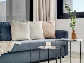 A modern living room with a grey sofa, beige and white cushions, a small round table with a glass and book, and a vase with green plants. - Sanders Premier Downtown - Full Building (Limassol)