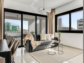 A modern living room with a grey sofa, a coffee table, and a view of the cityscape through large windows. - Sanders Premier Downtown - Full Building (Limassol)