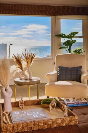 Een woonkamer met uitzicht op zee, een beige fauteuil, een rond bijzettafeltje en decoratieve objecten, waaronder een vaas met pluimveedergras en een klein sculptuur.