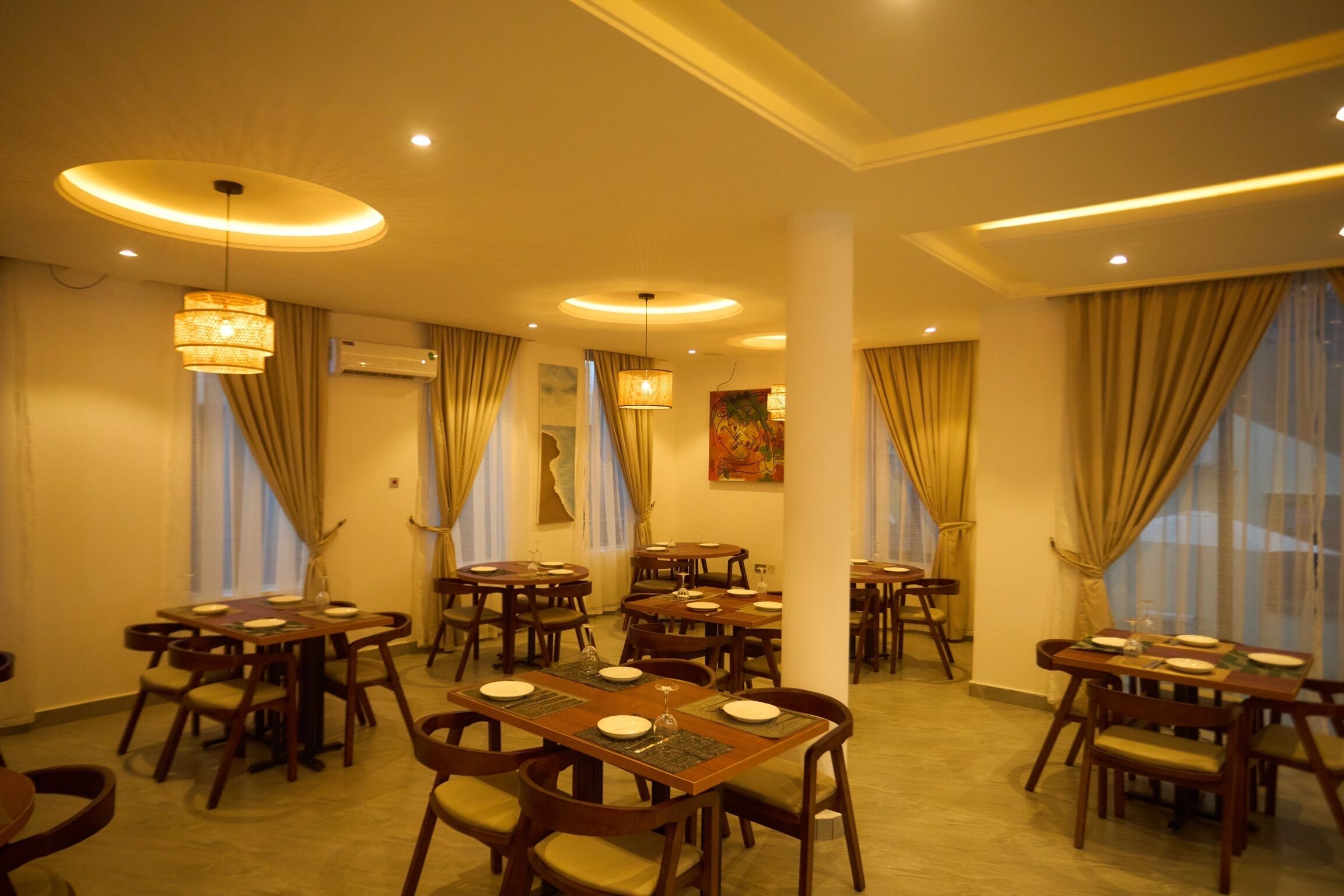 A hotel dining area with multiple tables and chairs, ceiling lights, and large windows with curtains.