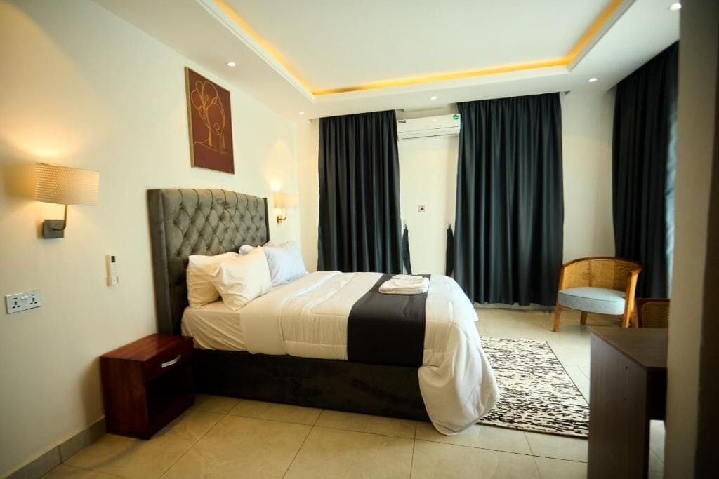 Habitación de hotel con una cama grande, una mesita de noche, una lámpara, una silla y una ventana con cortinas.