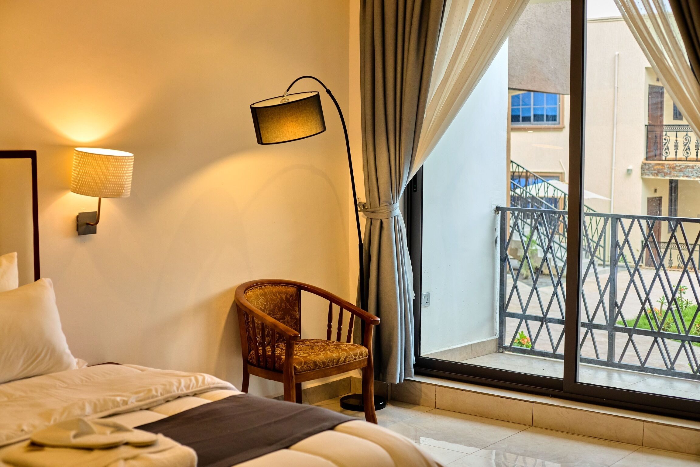 A hotel room with a bed, a chair, a lamp, and a view of the outside balcony.