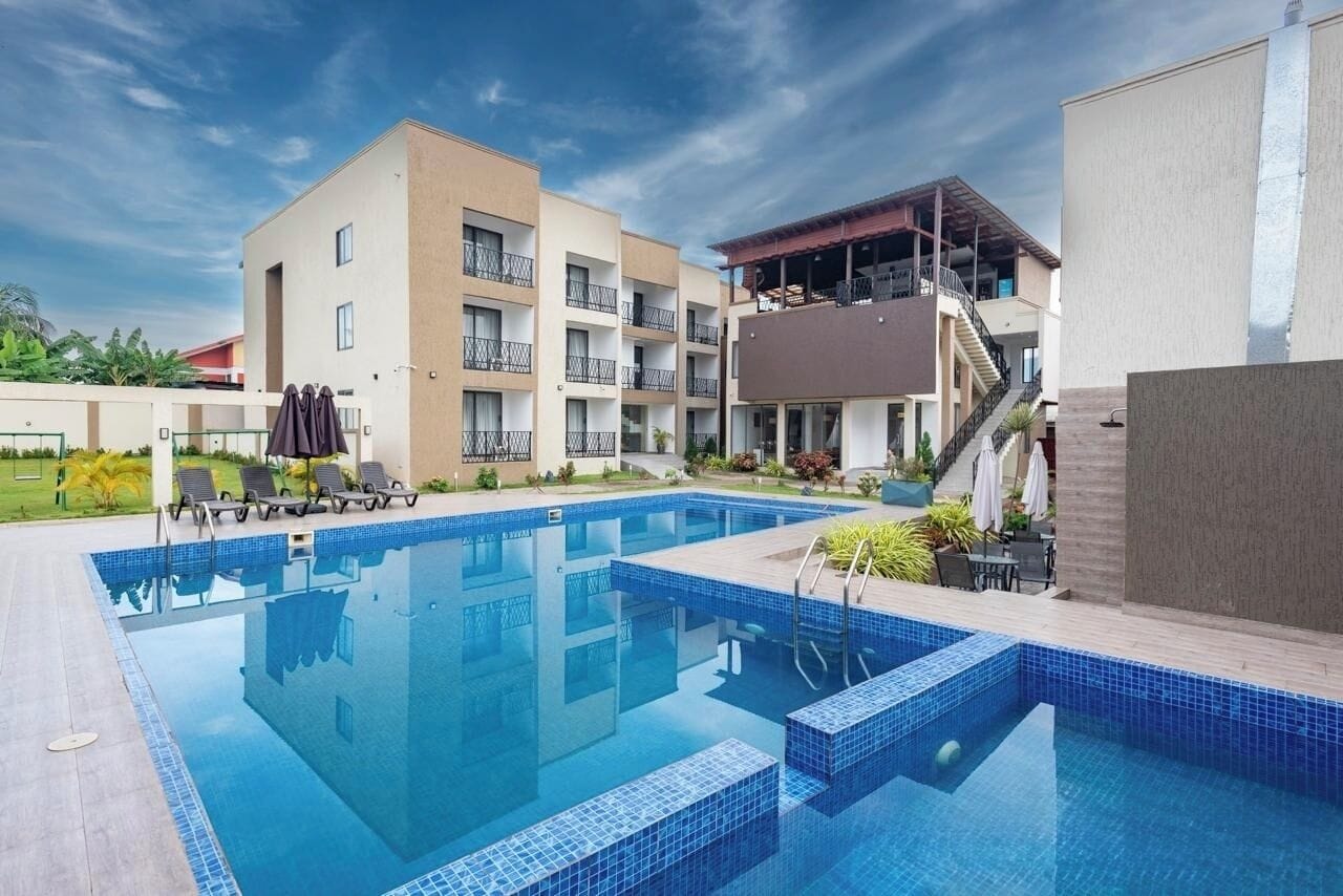 A swimming pool with lounge chairs and a covered area, surrounded by buildings with balconies and a staircase.
