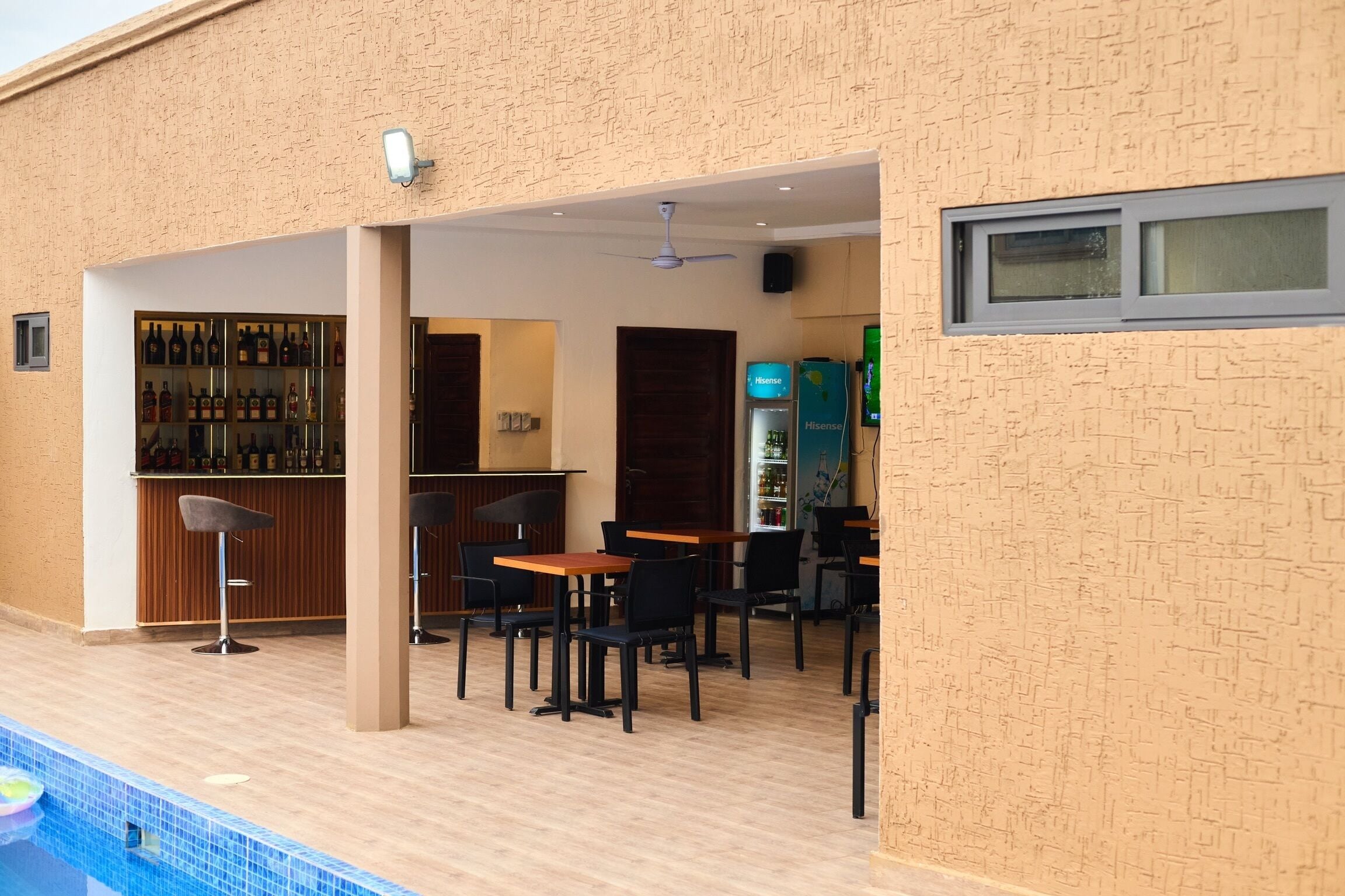 Angolo bar con varie bottiglie, zona salotto con tavoli e sedie e vista sulla piscina.