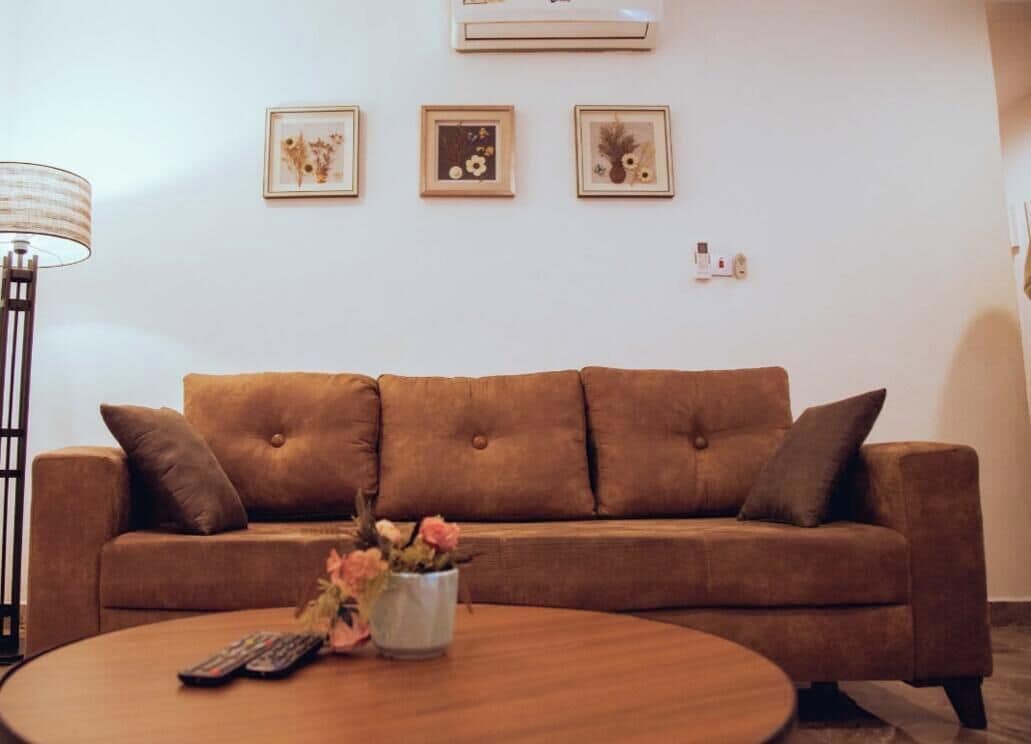 A living room with a brown sofa, a coffee table with a vase of flowers, a floor lamp, and three framed pictures on the wall.