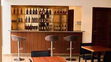 Una zona de bar con varias estanterías llenas de botellas, tres taburetes y una mesa de madera con sillas.