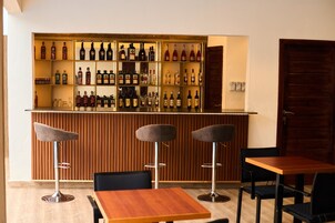 Una zona de bar con varias estanterías llenas de botellas, tres taburetes y una mesa de madera con sillas.