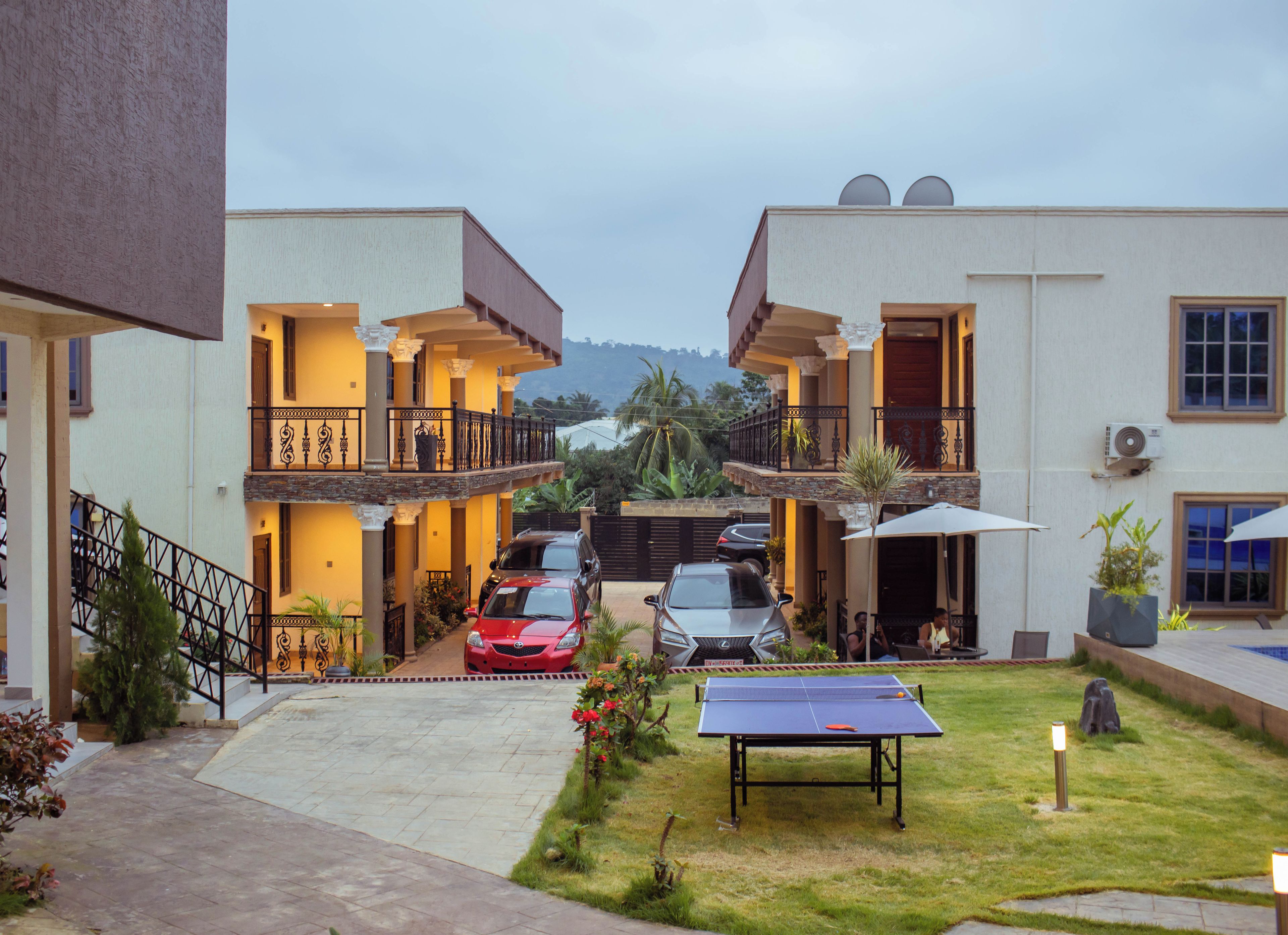Um edifício residencial com área externa coberta, mesa de tênis e carros estacionados.
