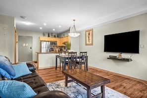 A modern living room with a wooden dining table, a leather sofa, and a flat-screen TV mounted on the wall.