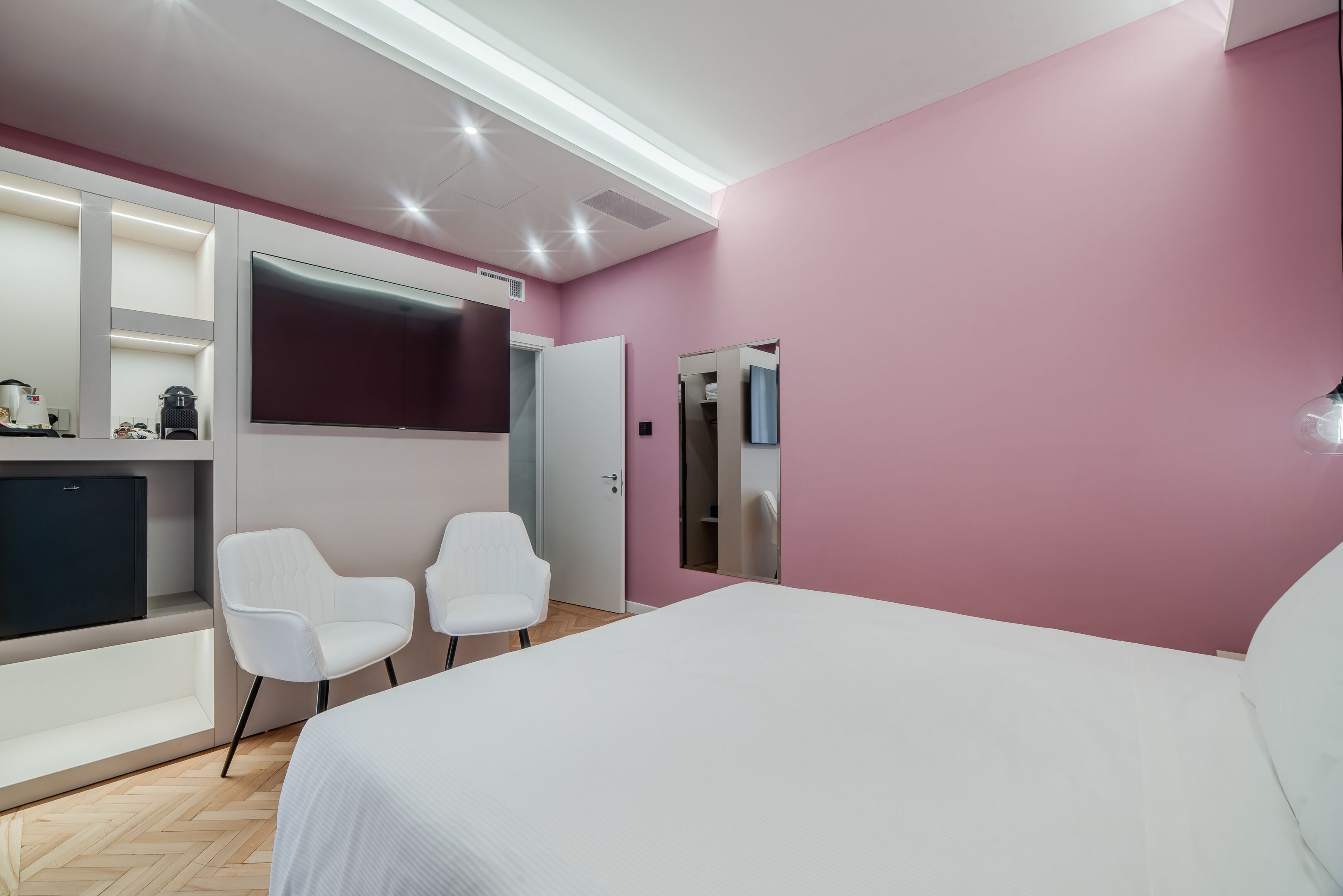 Camera d'albergo moderna con un letto grande, due poltrone bianche, una televisione a schermo piatto, un mini-frigo e un armadio.