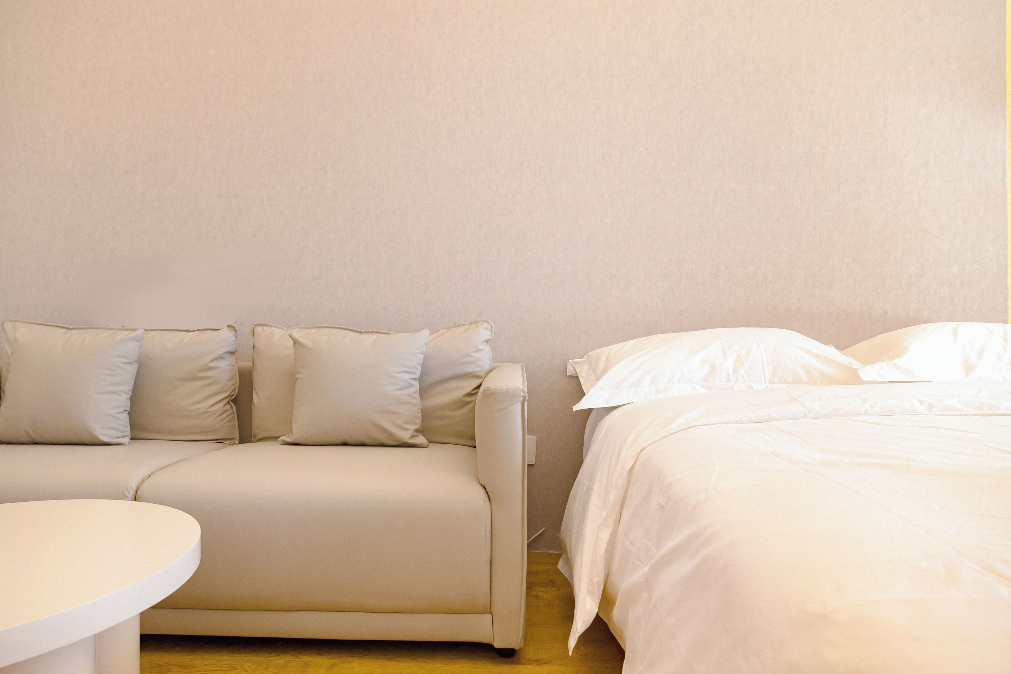 A room with a beige sofa, a round white table, and a bed with white bedding.