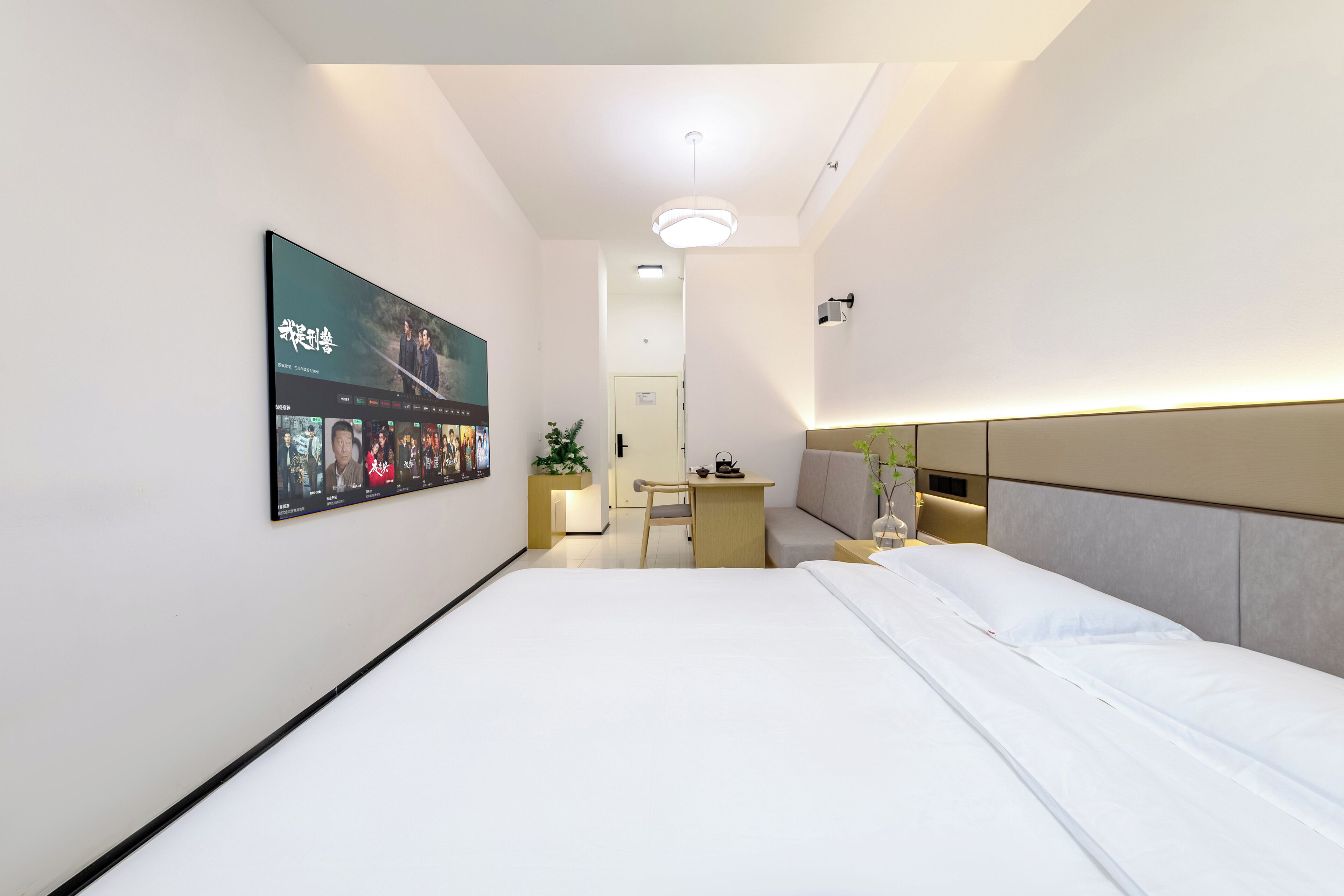 A modern hotel room with a large bed, a desk, and a TV mounted on the wall displaying a movie.