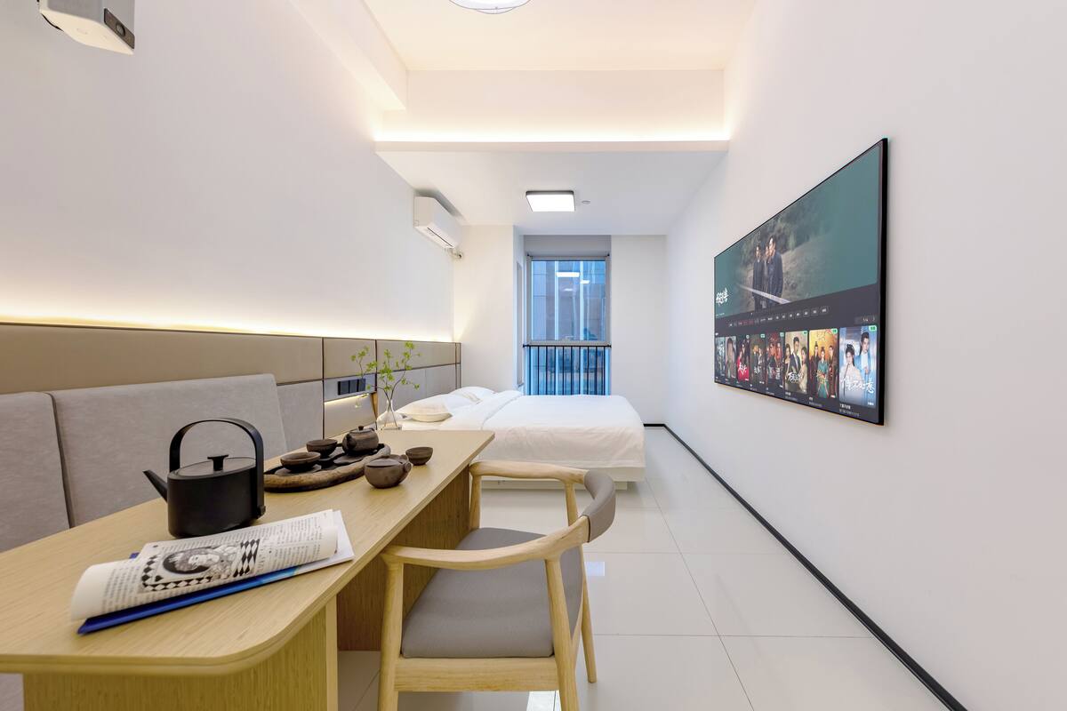 Ett modernt hotellrum med en säng, ett skrivbord med en vattenkokare, en stol och en stor TV-skärm som visar en film.