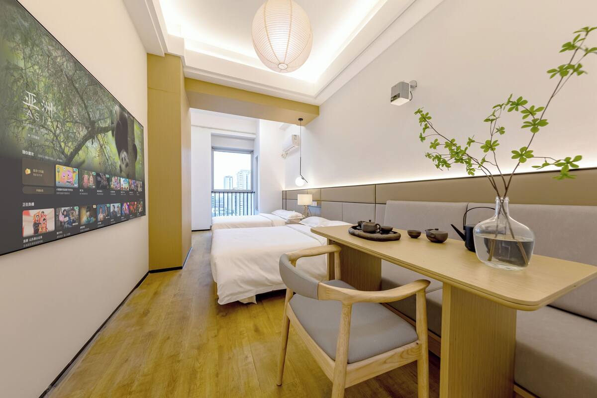Ett modernt hotellrum med trägolv, en säng med vita sängkläder, ett litet bord med en vas med gröna växter och en stor affisch på väggen.