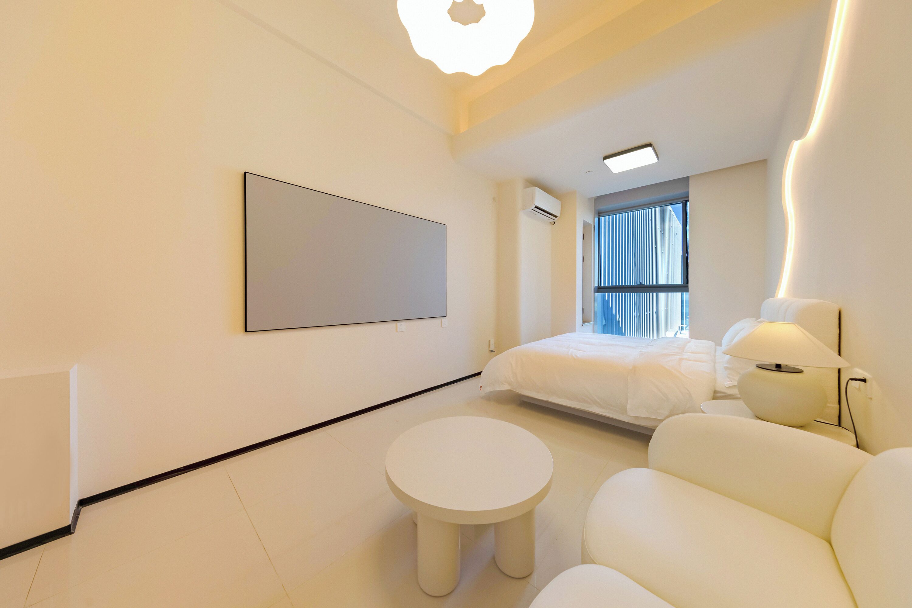 A modern hotel room with a large bed, a round white table, a sofa, a wall-mounted TV, and a window with curtains.