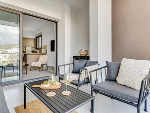 Un balcone moderno con una porta vetrata che dà su un soggiorno, un divano, un tavolino con bicchieri da vino e vista sulla città.