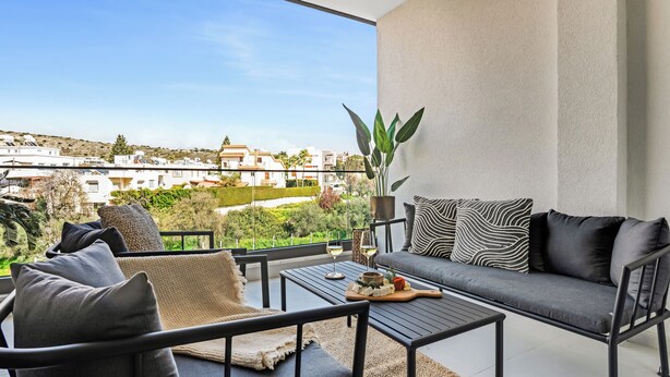 Un balcone con divano, sedie e tavolo, affaccia su una zona residenziale con case e vegetazione.