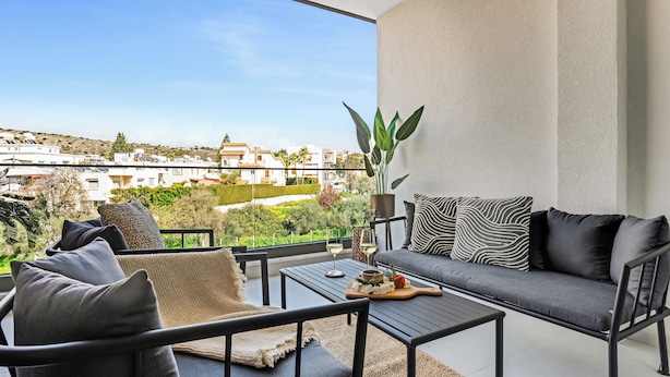 Un balcone con divano, sedie e tavolo, affaccia su una zona residenziale con case e vegetazione.