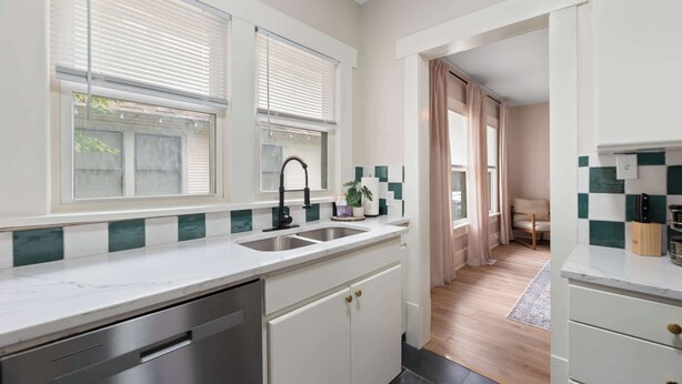 A modern kitchen with a white countertop, stainless steel sink, and a dishwasher. There are two windows with blinds, a potted plant, and a checkered backsplash.