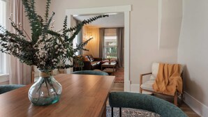 A dining area with a wooden table, chairs, and a vase with flowers. There is a sofa and a chair in the background.