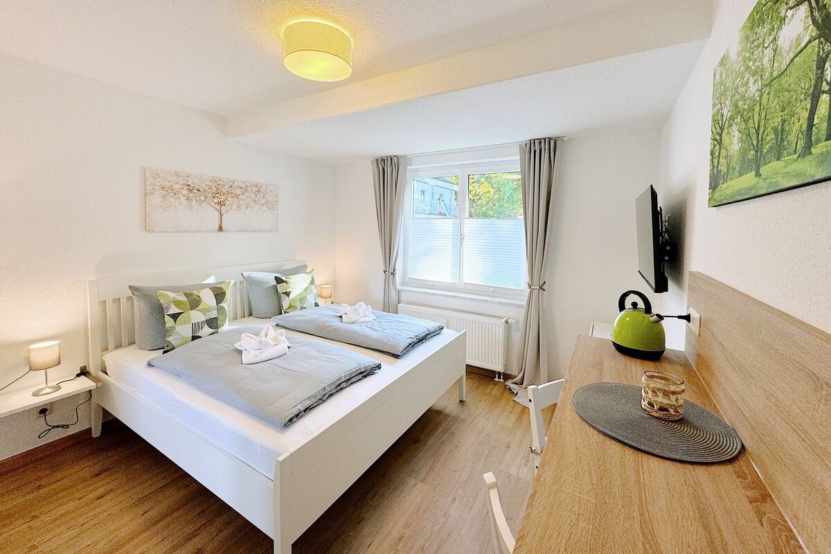 A modern bedroom with a large bed, bedside tables, a window with curtains, a painting, and a green kettle on the sideboard.