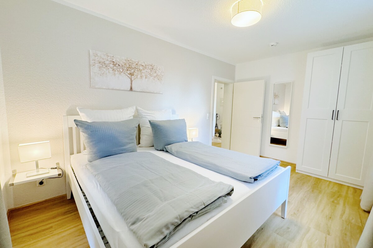 Un dormitorio con dos camas, paredes blancas, una lámpara en el techo y una puerta que da a otra habitación.