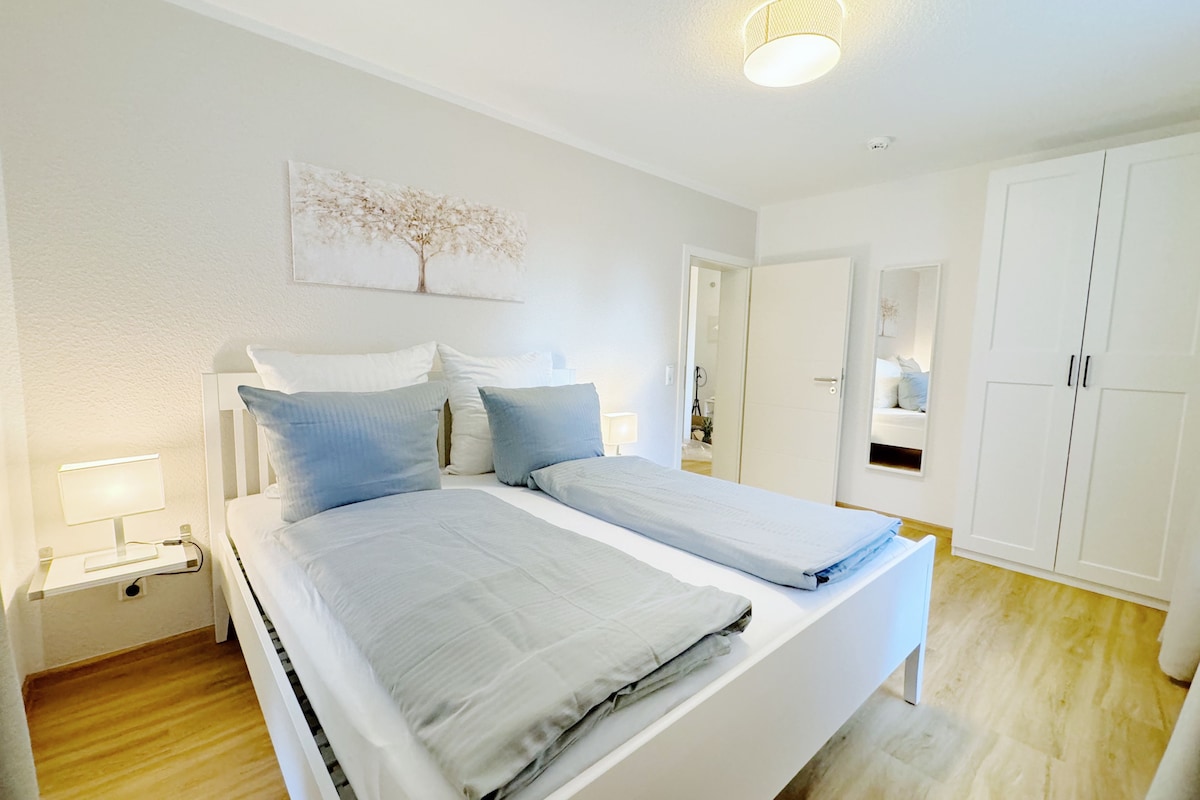 Un dormitorio con dos camas, paredes blancas, una lámpara en el techo y una puerta que da a otra habitación.
