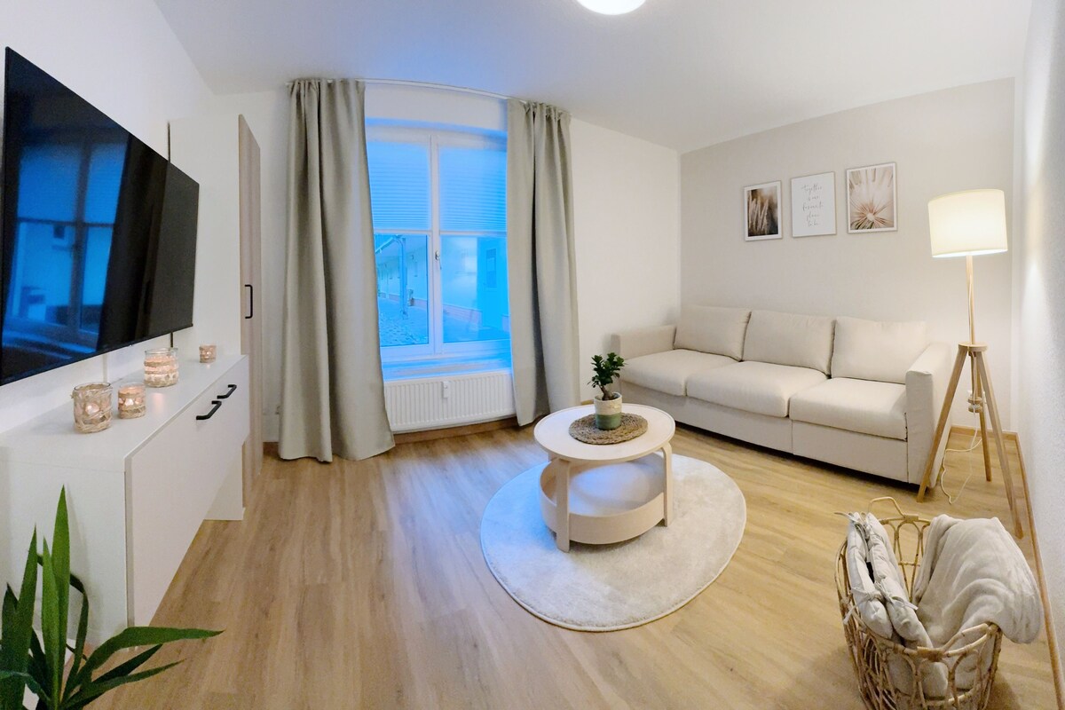 A modern living room with a flat-screen TV, a white sofa, a round coffee table, and a window with curtains.