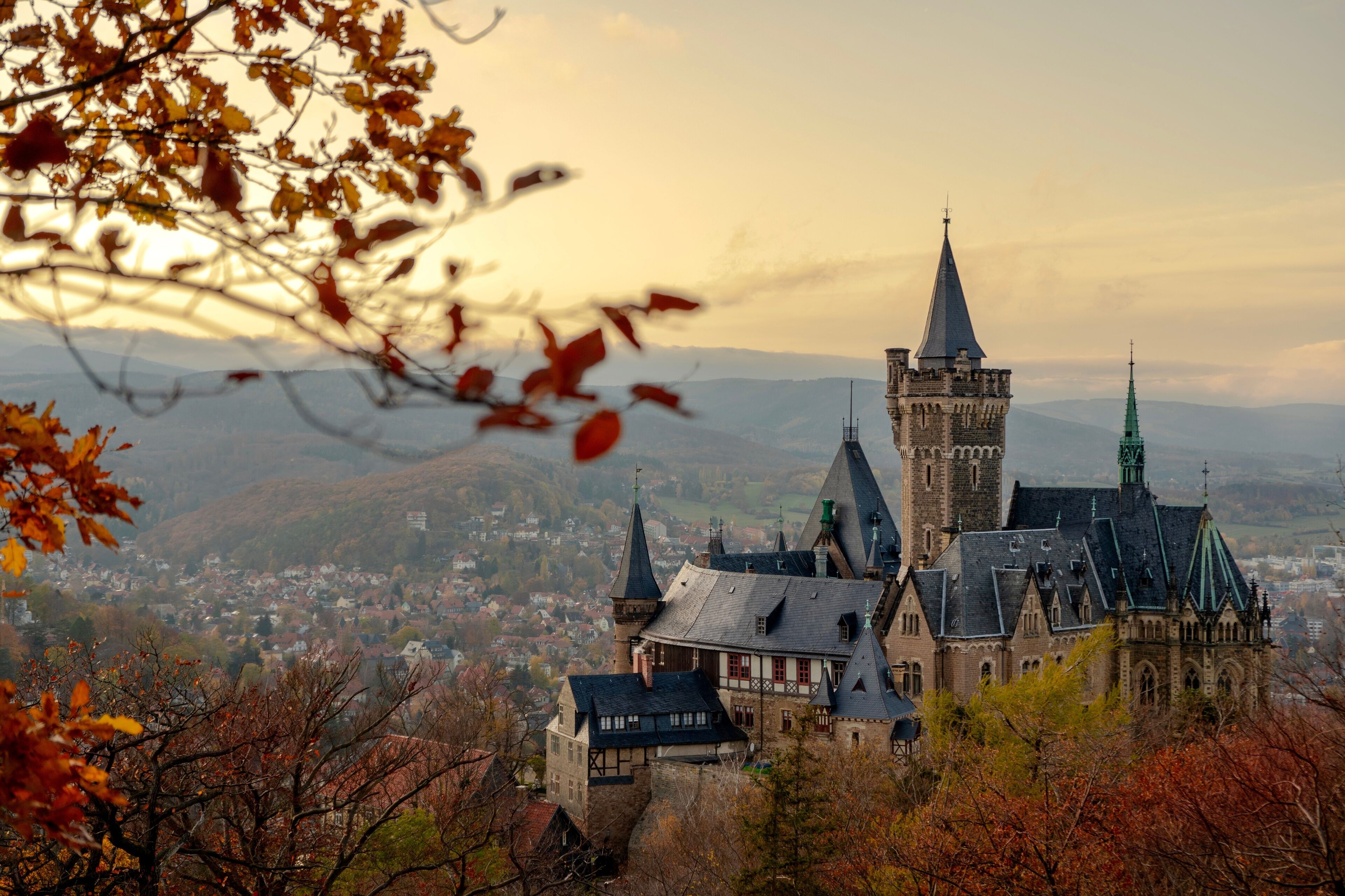A historic castle with multiple towers and a prominent central tower, surrounded by autumn foliage and a view of a town below.