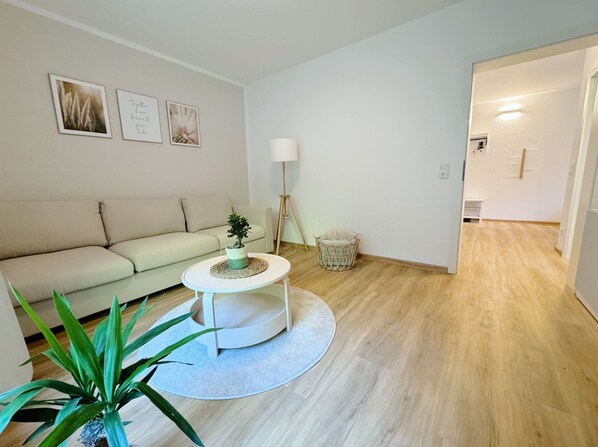 Una sala de estar moderna con un sofá color beige, una mesa redonda de centro con una planta, una lámpara de pie y una pequeña cesta de mimbre.