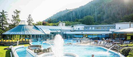 Une piscine avec une fontaine, entourée de chaises longues et bordée d’un bâtiment à la façade en verre.