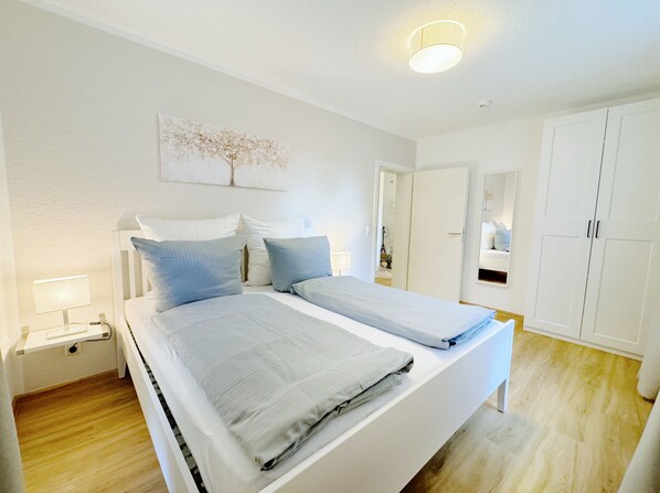 Un dormitorio con dos camas, paredes blancas, una lámpara en el techo y una puerta que da a otra habitación.