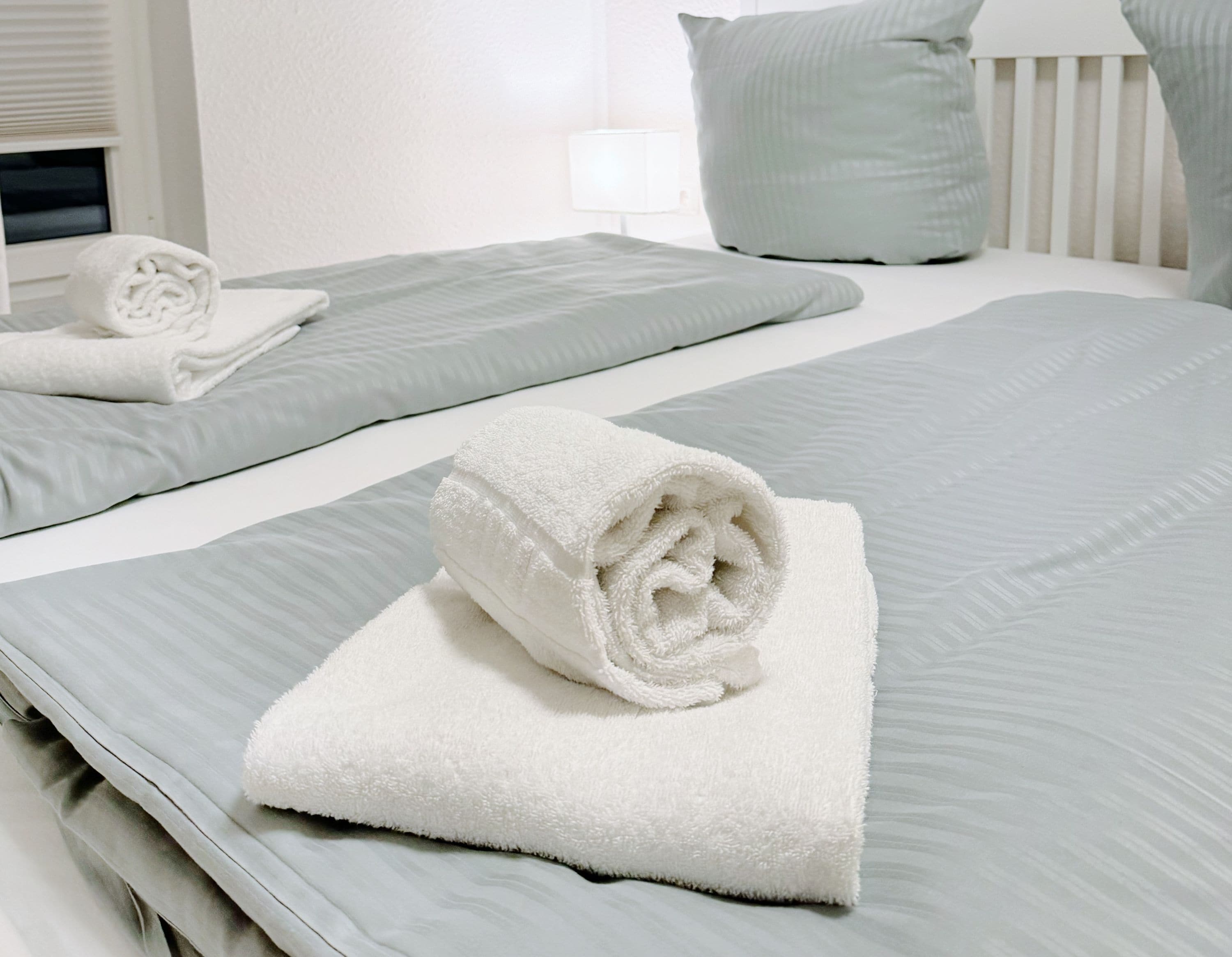 A neatly made bed with a rolled white towel, a grey bedspread, and a matching pillow.