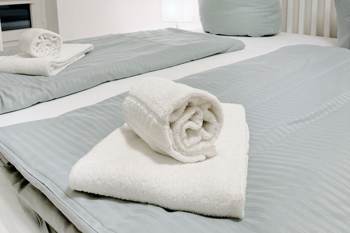 A neatly made bed with a rolled white towel on top, light blue bedding, and a white pillow.