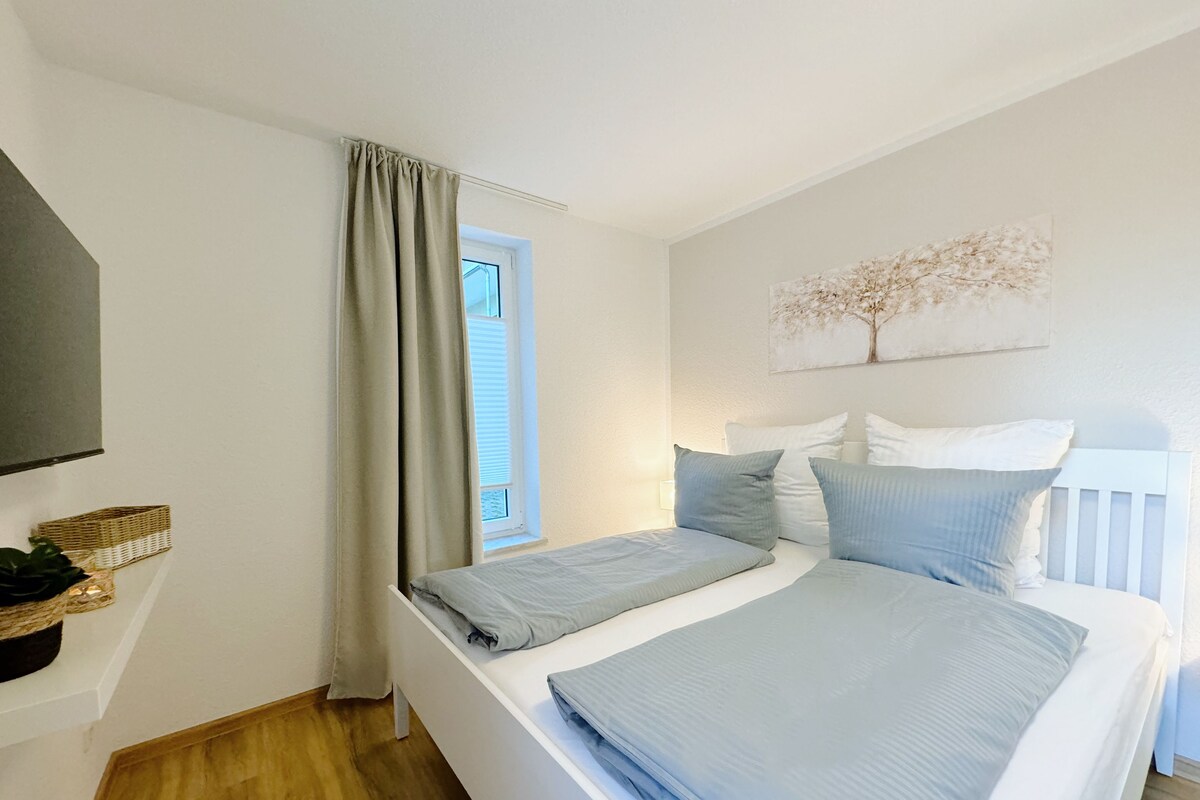 Un dormitorio con cama, ventana con cortinas, televisor de pared y un cuadro de un árbol.