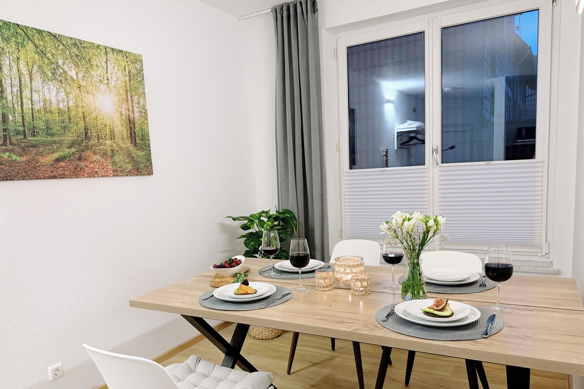A dining area with a wooden table set for four, surrounded by white chairs. There are plates with food, glasses of red wine, and a vase with flowers. A large window with blinds is visible, and a painting of a forest hangs on the wall.