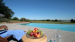 A poolside setting with a wicker basket of fruit and glasses of water, a wooden chair, and a clear view of a swimming pool and landscape.