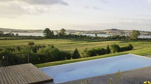 A swimming pool with a wooden deck, surrounded by a grassy landscape and trees, overlooking a body of water and hills. - POGGIO CASALE (Chiusi)
