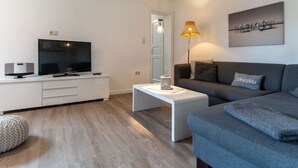 En moderne stue med et fladskærms-tv, et hvidt sofabord og en grå sofa samt en lampe.