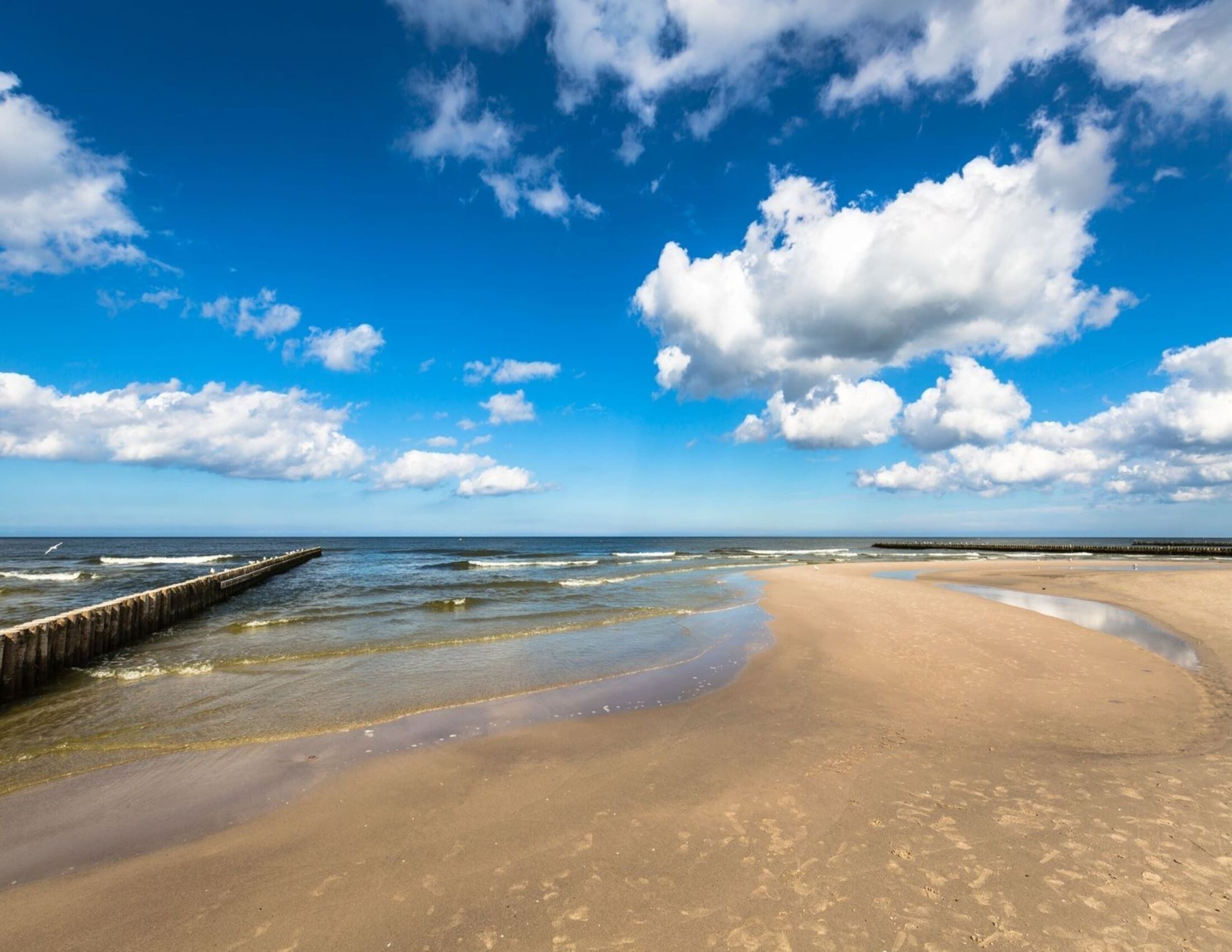 Una playa con un muelle de madera que se adentra en el agua, orilla arenosa y un cielo azul despejado con algunas nubes.