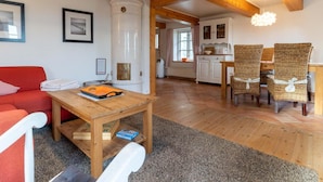 A cozy living room with a red sofa, wooden coffee table, and wicker chairs. A kitchen with white cabinets and a window is visible in the background. - Ferienhaus in Westerhever mit Garten (Westerhever)