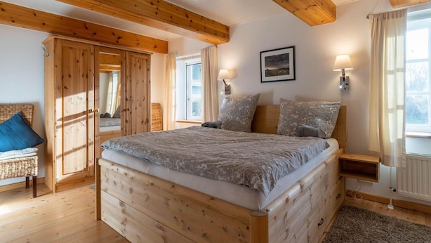 A bedroom with a wooden bed, a nightstand, a lamp, and a window with curtains. - Ferienhaus in Westerhever mit Garten (Westerhever)