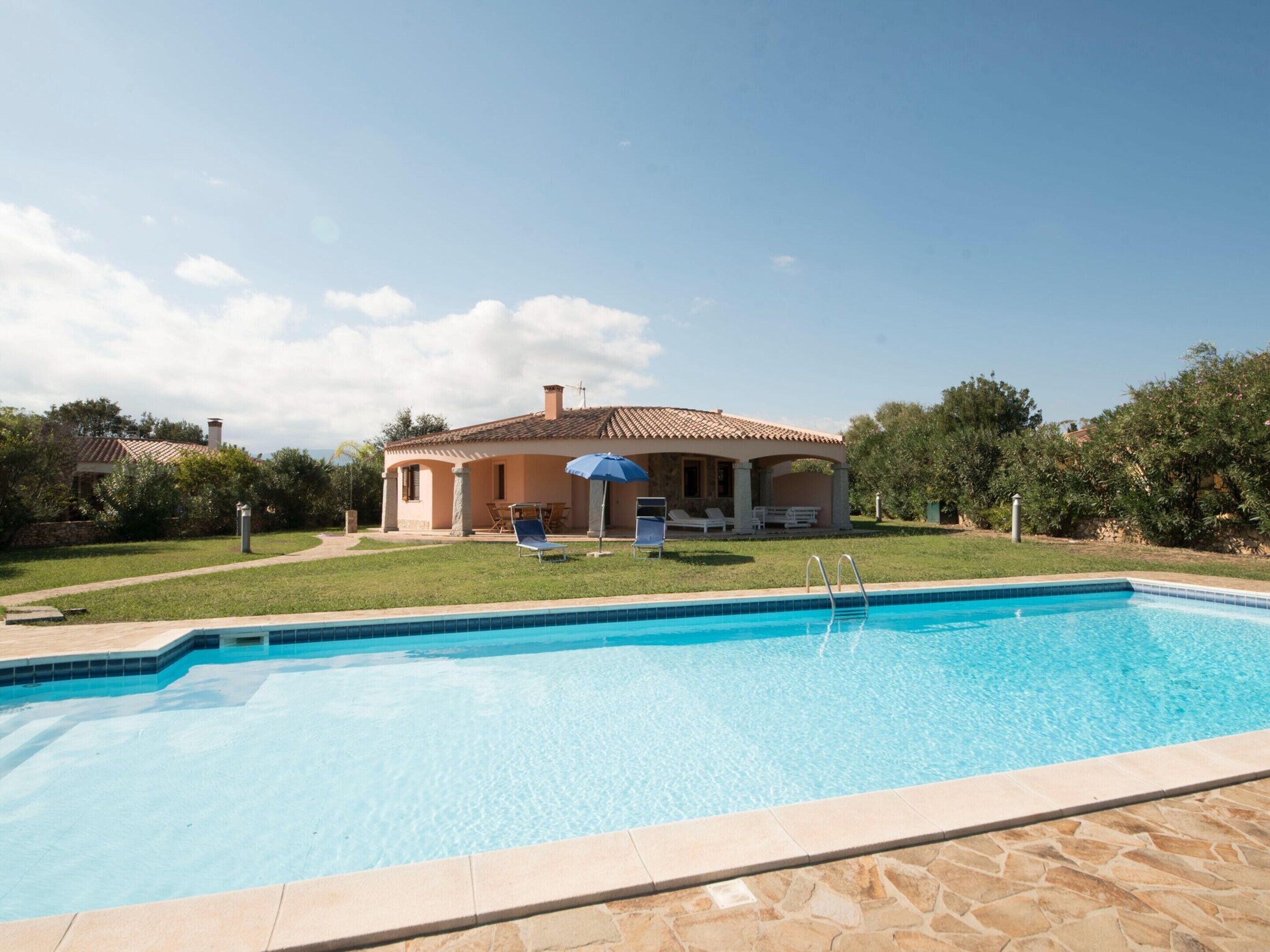 A swimming pool with a stone border, a house with a tiled roof, and a lawn with a blue umbrella.