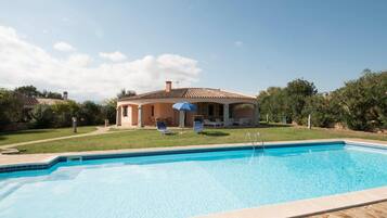 A swimming pool with a stone border, a house with a tiled roof, and a lawn with a blue umbrella.