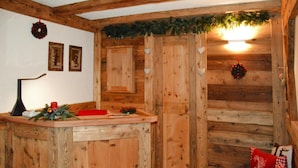 A rustic wooden cabin interior with a wooden bar counter, framed pictures on the wall, and a cozy seating area with red and white cushions. - Apartment in Pellizzano Near Shopping (Pellizzano)