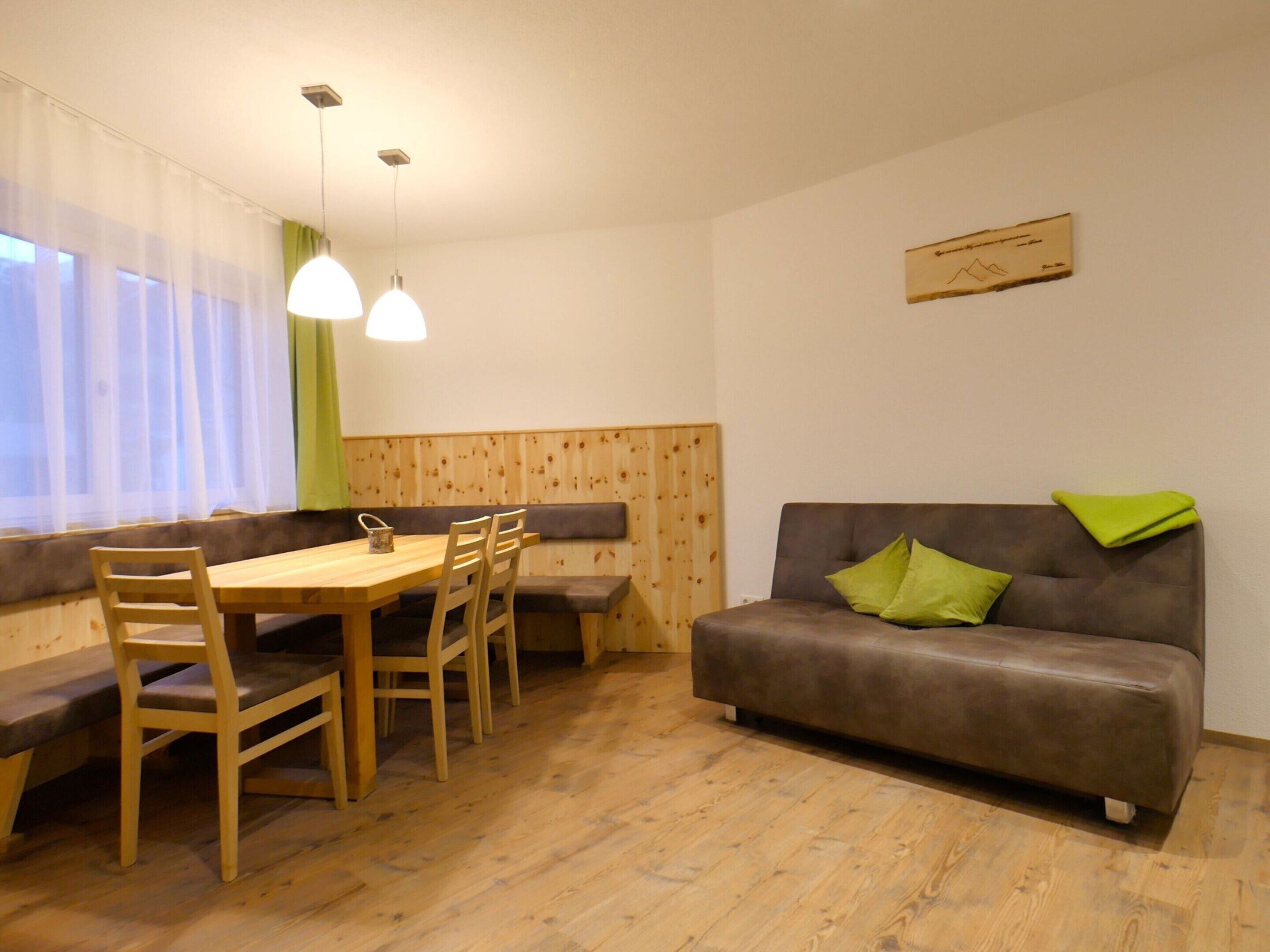 A room with a wooden dining table, four chairs, a bench, a brown sofa with green cushions, and a wooden wall panel.